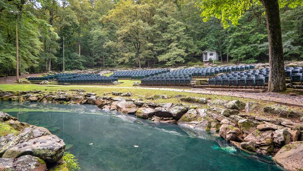 A natural setting with a clear pond surrounded by rocks and trees, and rows of blue chairs arranged for an event. - Pine Mountain State Resort Park (Pineville)