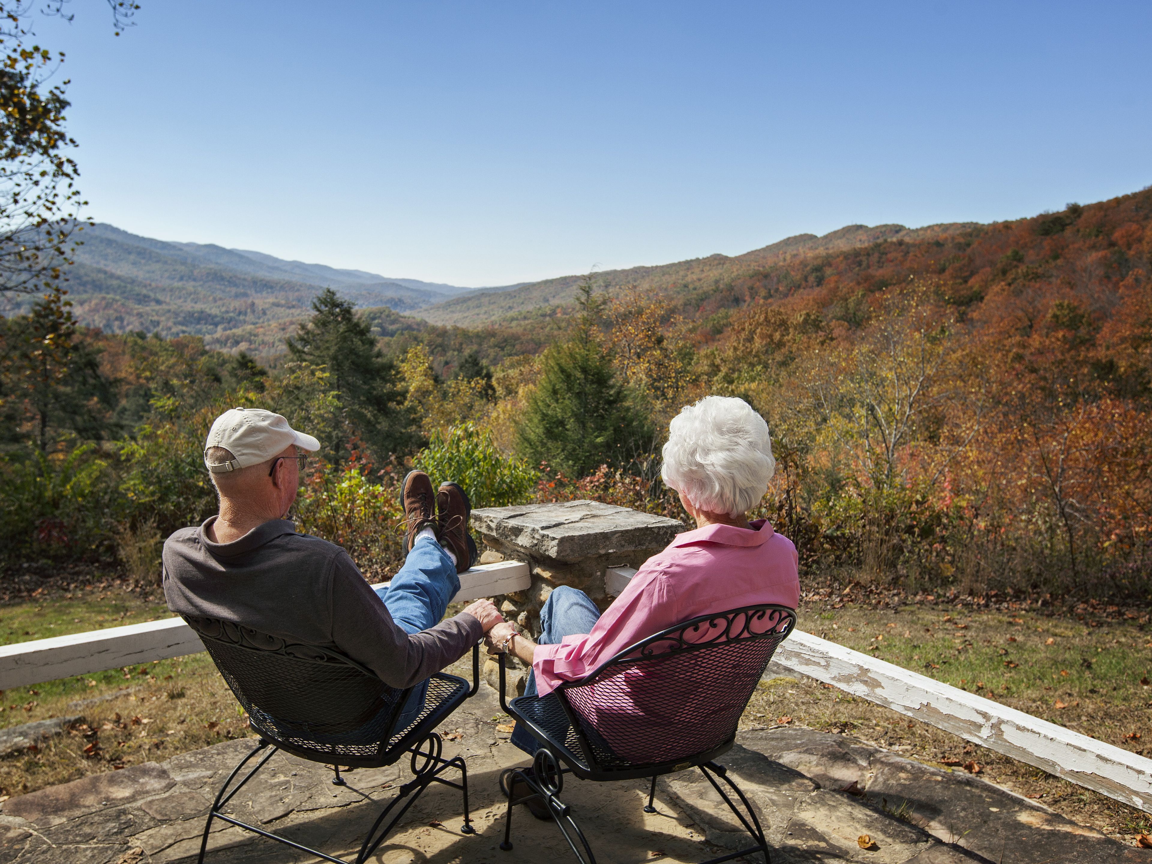 Três pessoas sentadas em cadeiras ao ar livre apreciando a vista panorâmica de uma paisagem montanhosa com folhagem de outono.