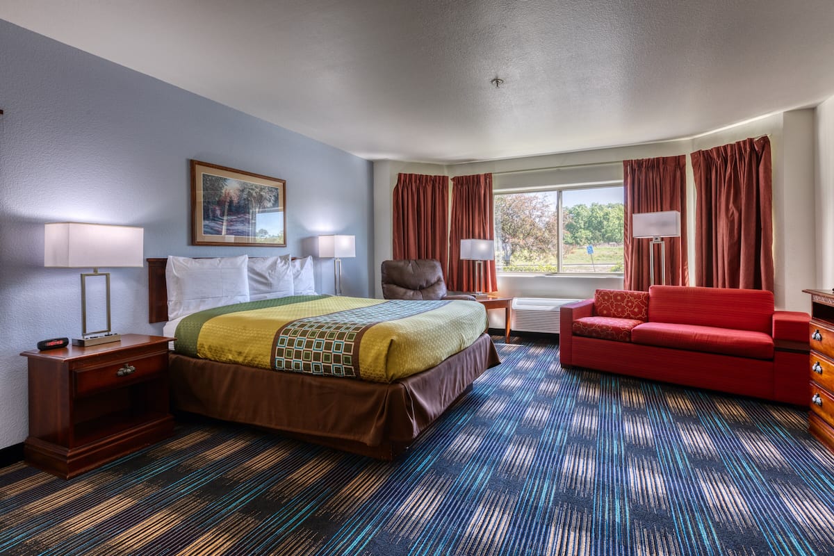 A hotel room with a bed, a red sofa, a nightstand, a lamp, and a window with curtains.
