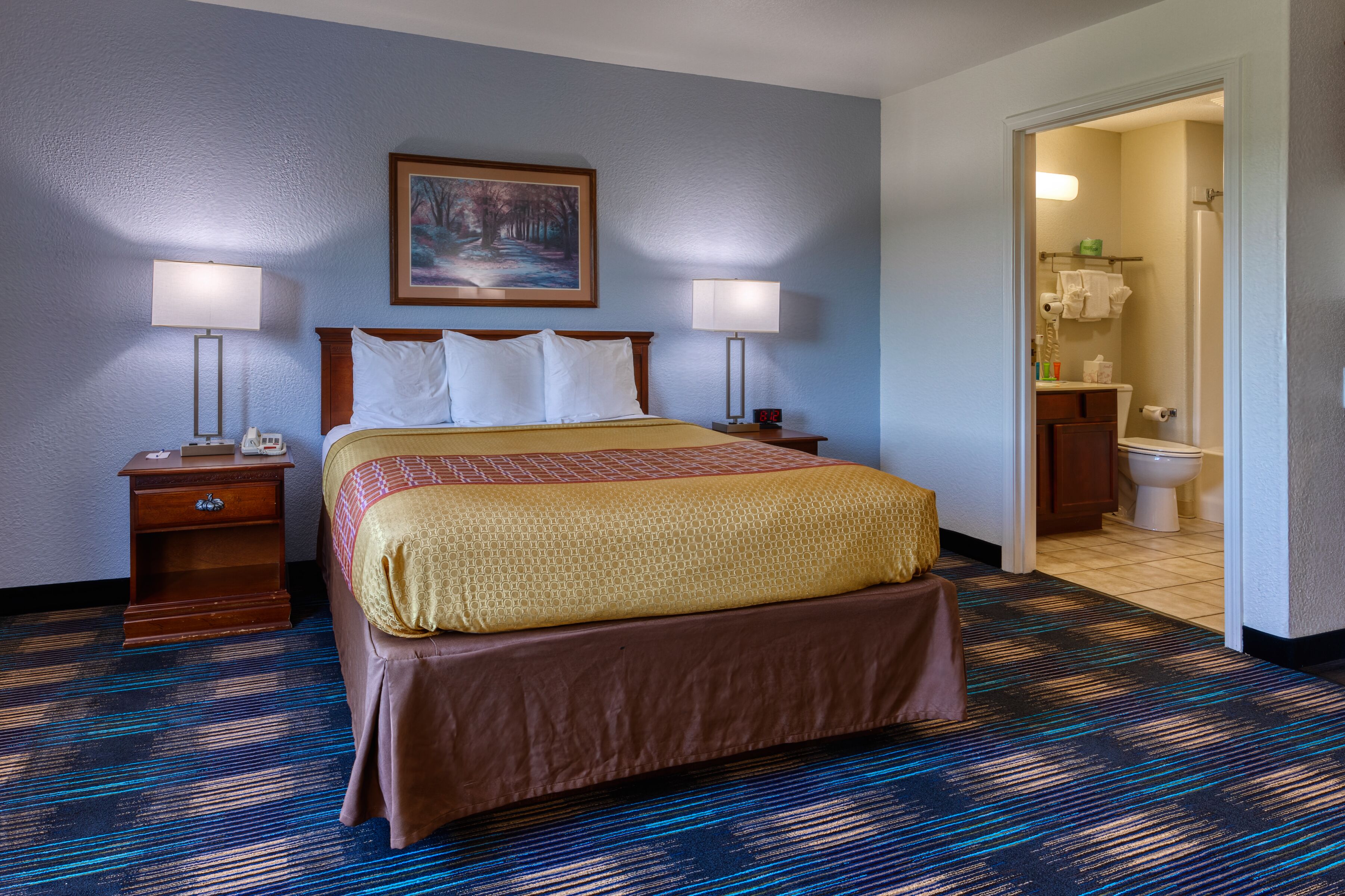 A hotel room with a bed, two bedside tables, a nightstand, a painting on the wall, and a bathroom visible through an open door.