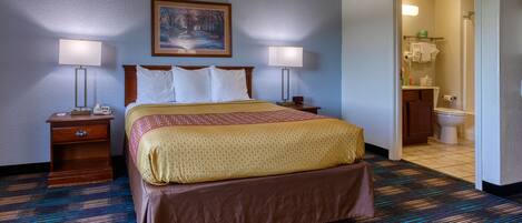 A hotel room with a bed, two bedside tables, a nightstand, a painting on the wall, and a bathroom visible through an open door.