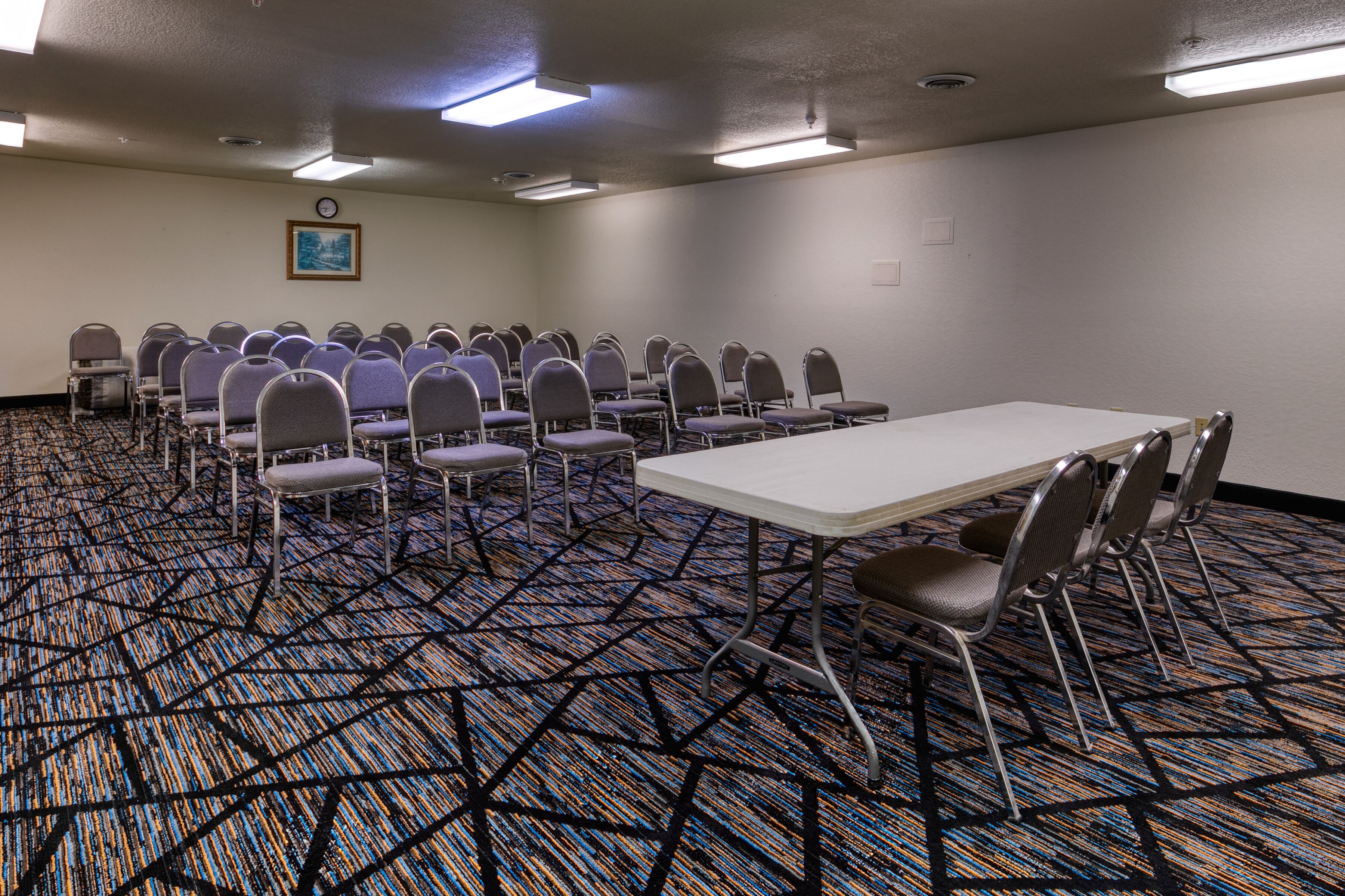 Un salón de conferencias con una larga mesa y filas de sillas, suelo alfombrado y un reloj en la pared.