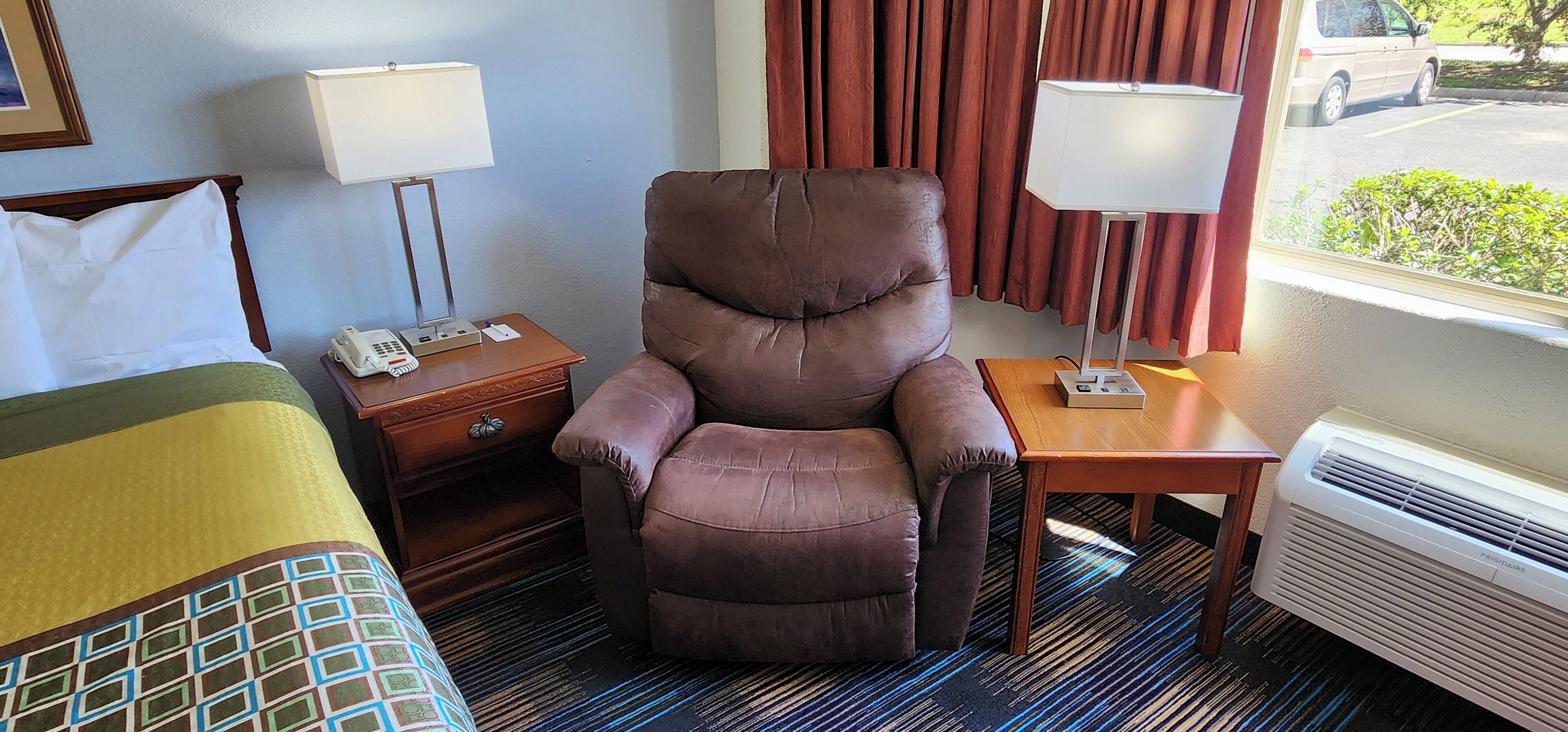 A hotel room with a large brown leather chair, a bed with a green and yellow patterned bedspread, two bedside tables with lamps, and a window with a view of a parking lot and greenery.
