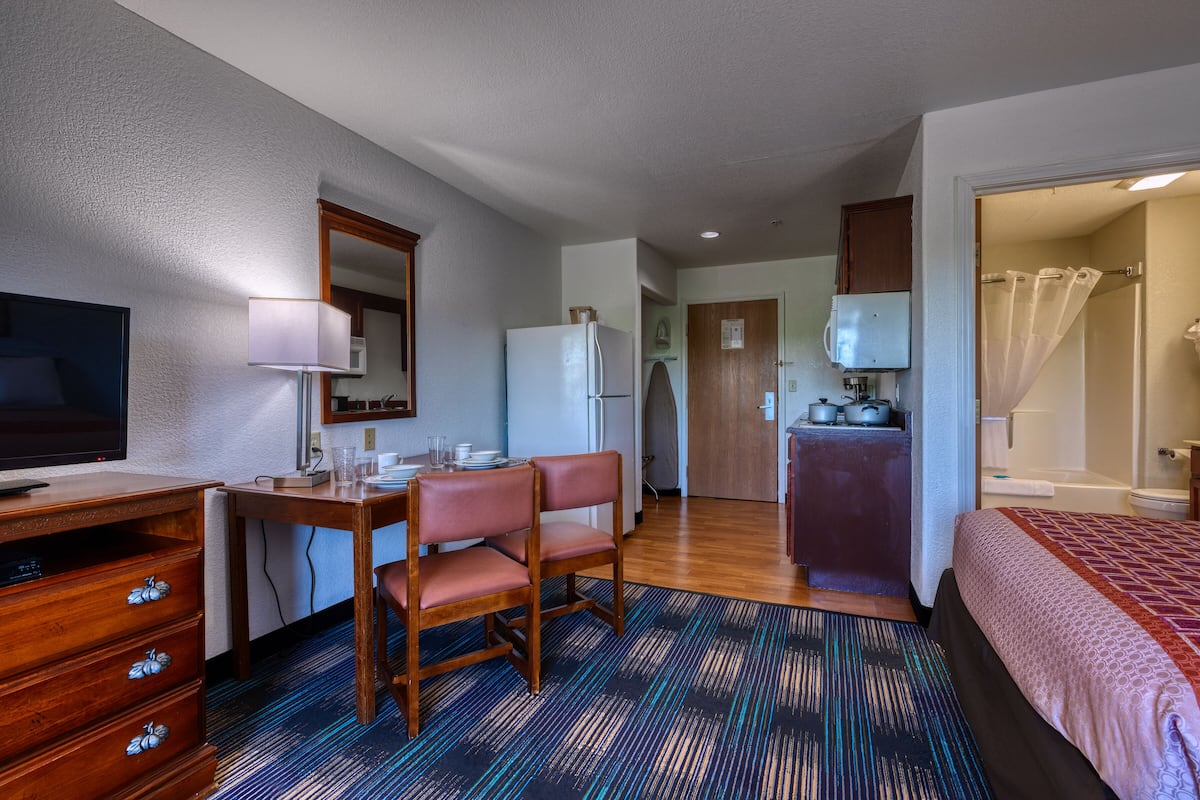 A compact hotel room with a bed, a desk with a chair, a television, a refrigerator, a microwave, and a bathroom with a shower curtain.