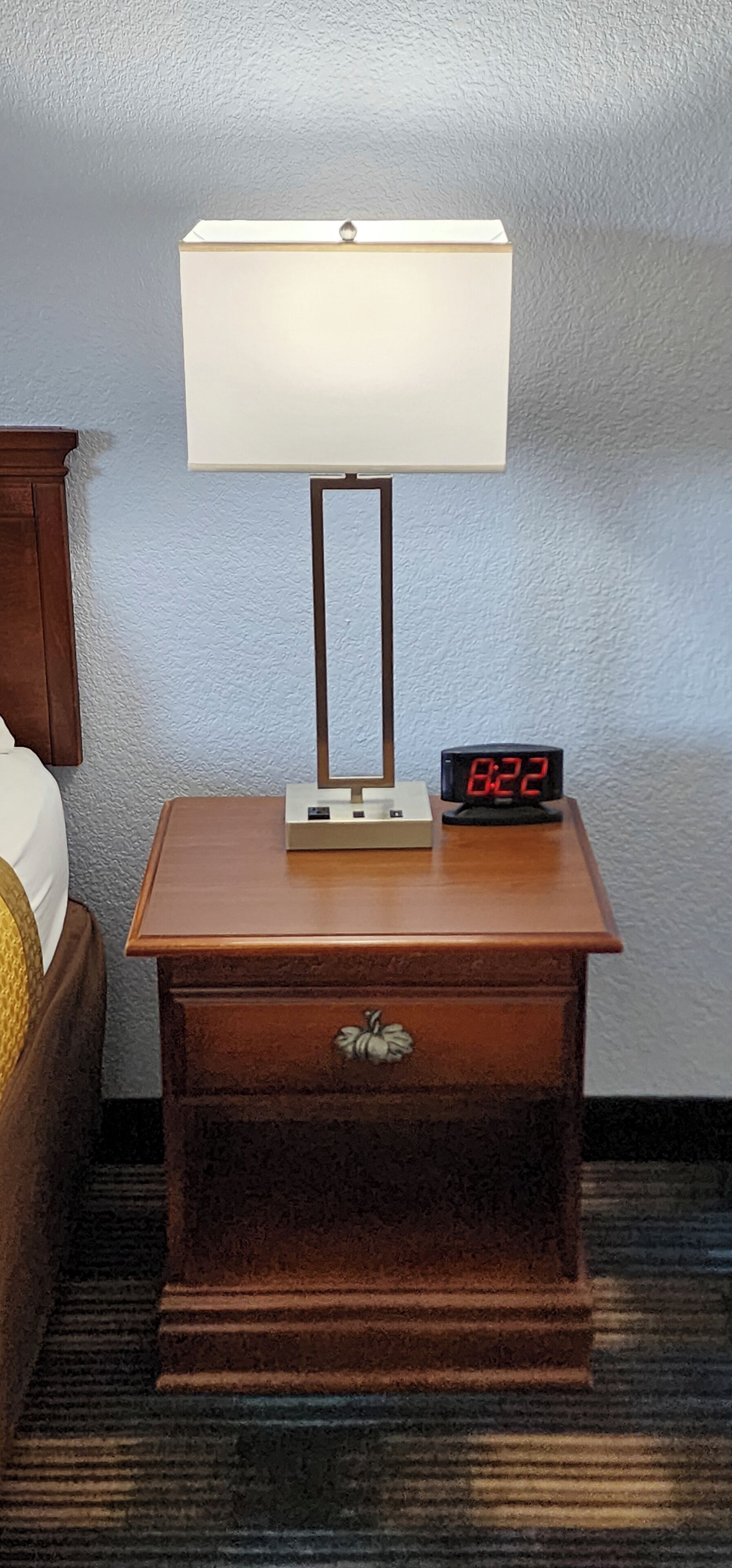 Una mesita de noche de madera con una lámpara blanca y un reloj digital que marca las 8:22.