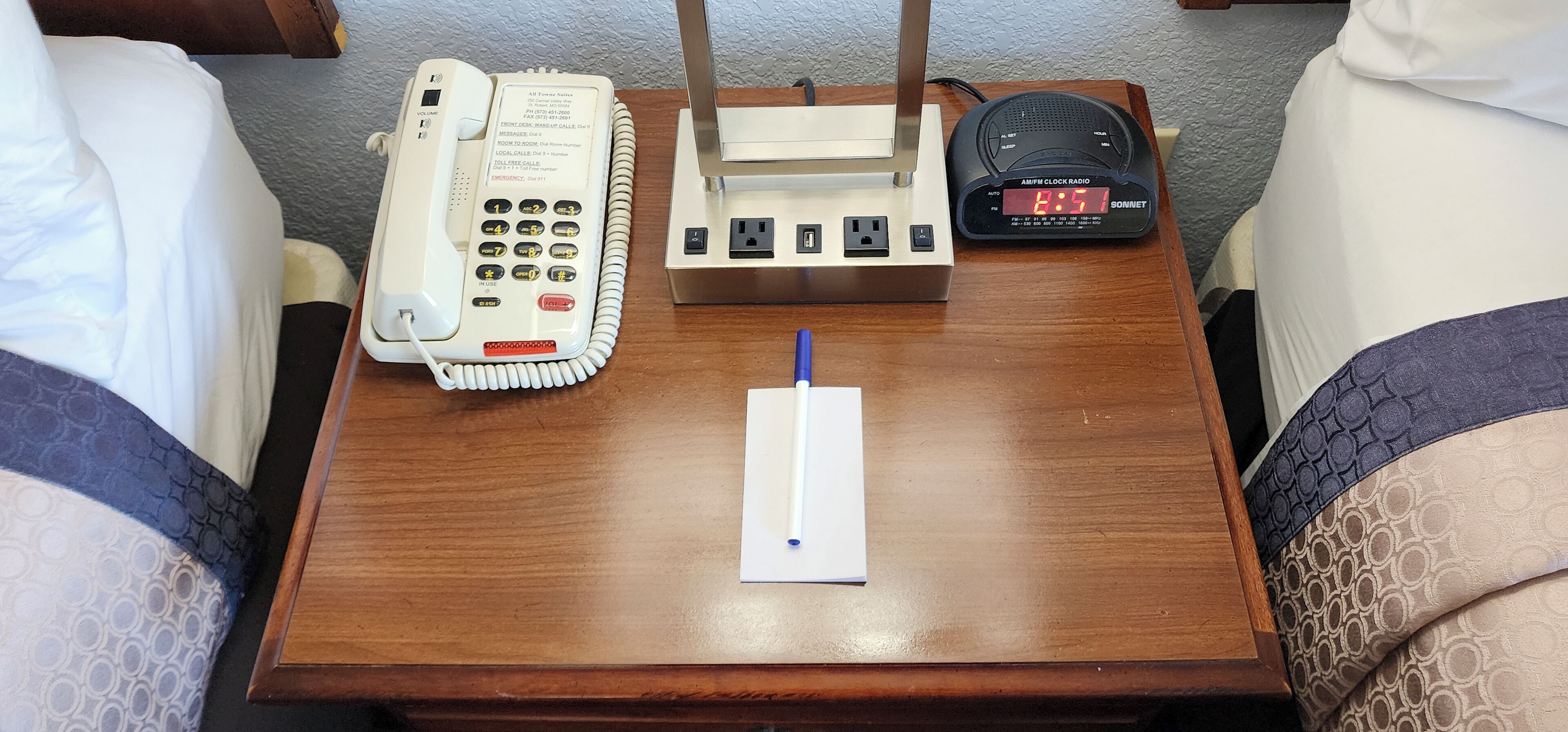 Una mesita de noche de madera con un teléfono blanco, una regleta y un reloj digital.