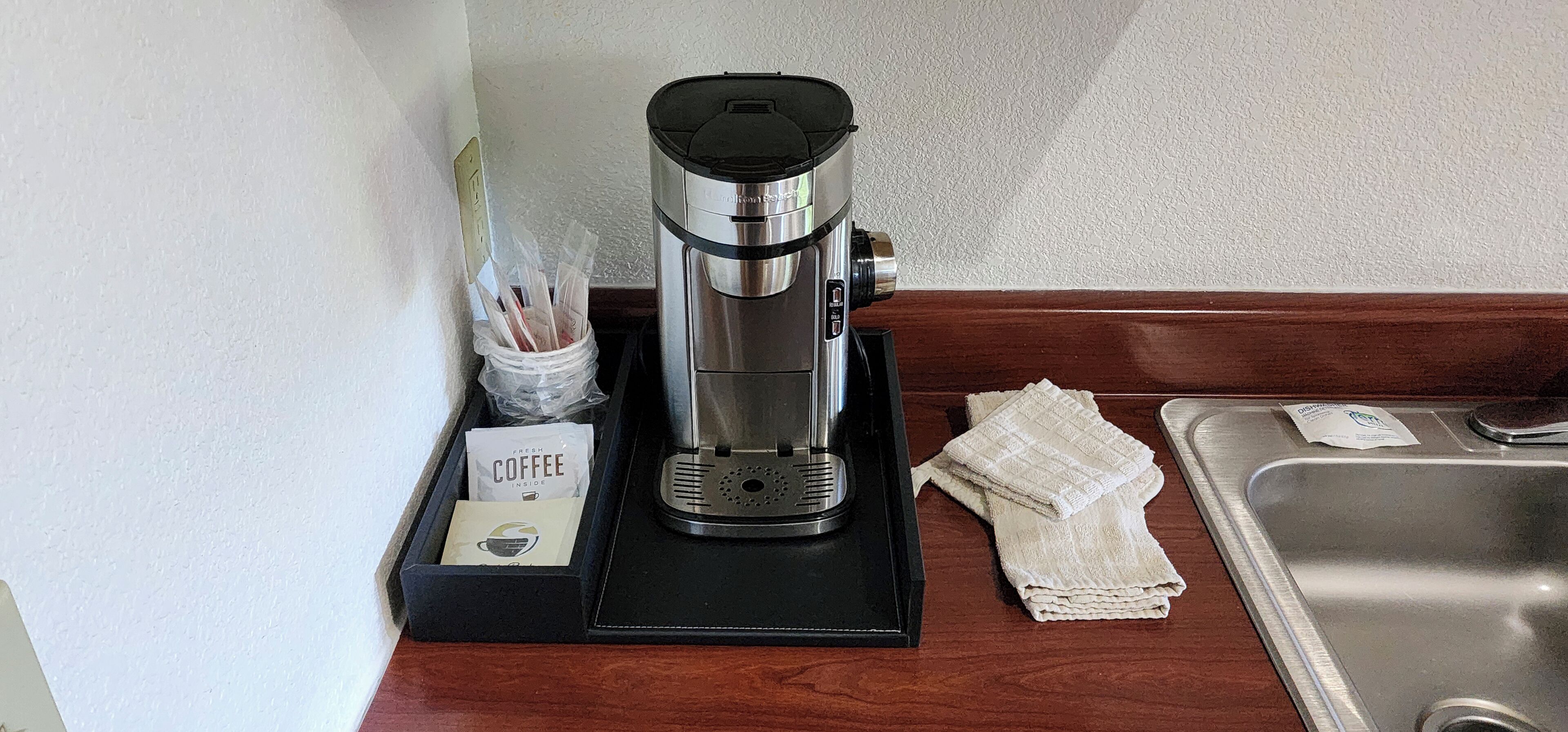 Een koffiezetapparaat op een zwarte dienblad, met een koffiekapsel en een zak koffiekapsels op het aanrecht.