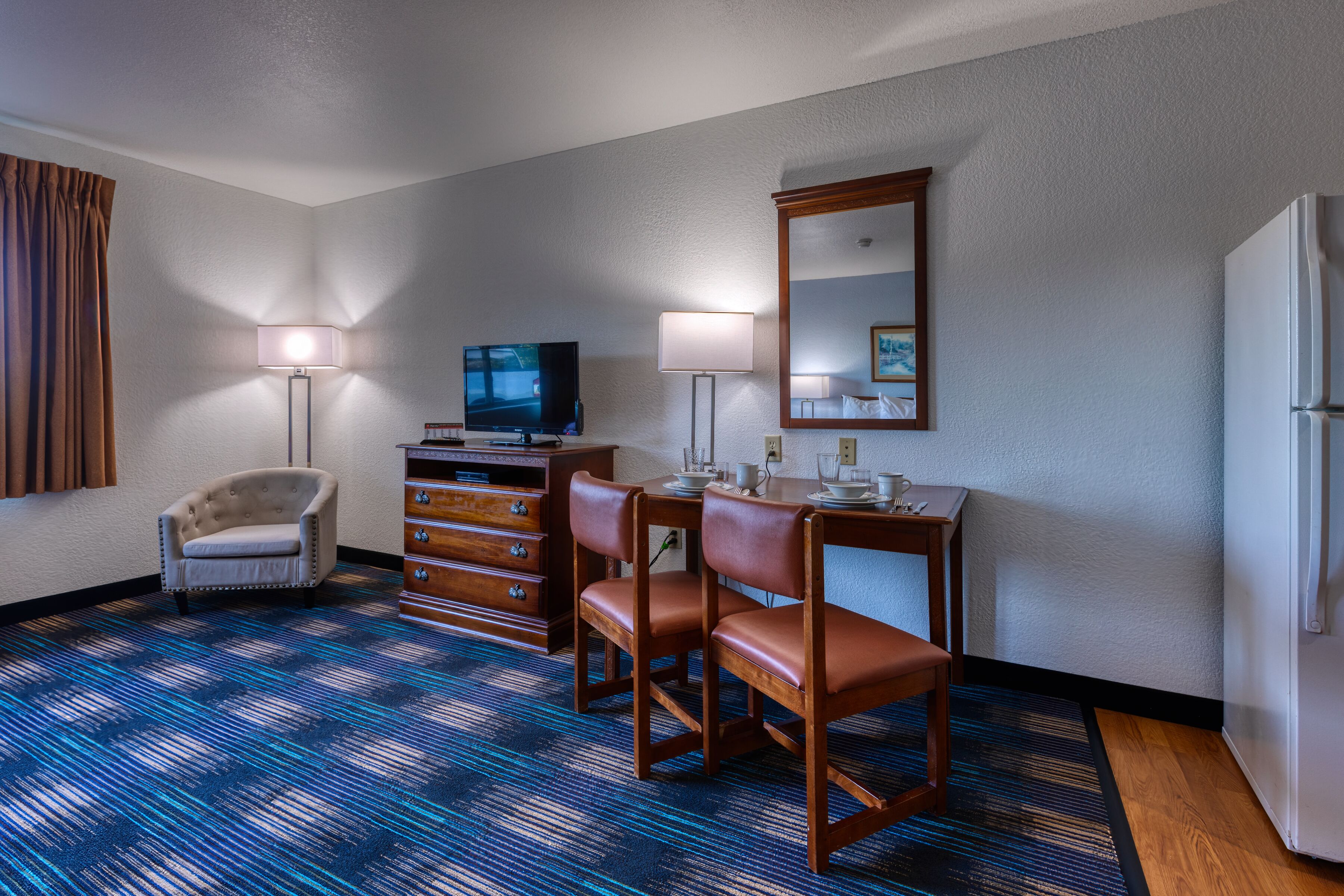 A hotel room with a blue patterned carpet, a wooden desk with a mirror, a television, a dresser, and a chair.