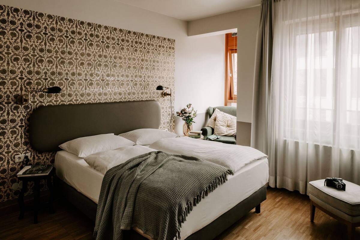 A neatly arranged bedroom with a large bed, a bedside table with a lamp, a small table with a book, and a chair.
