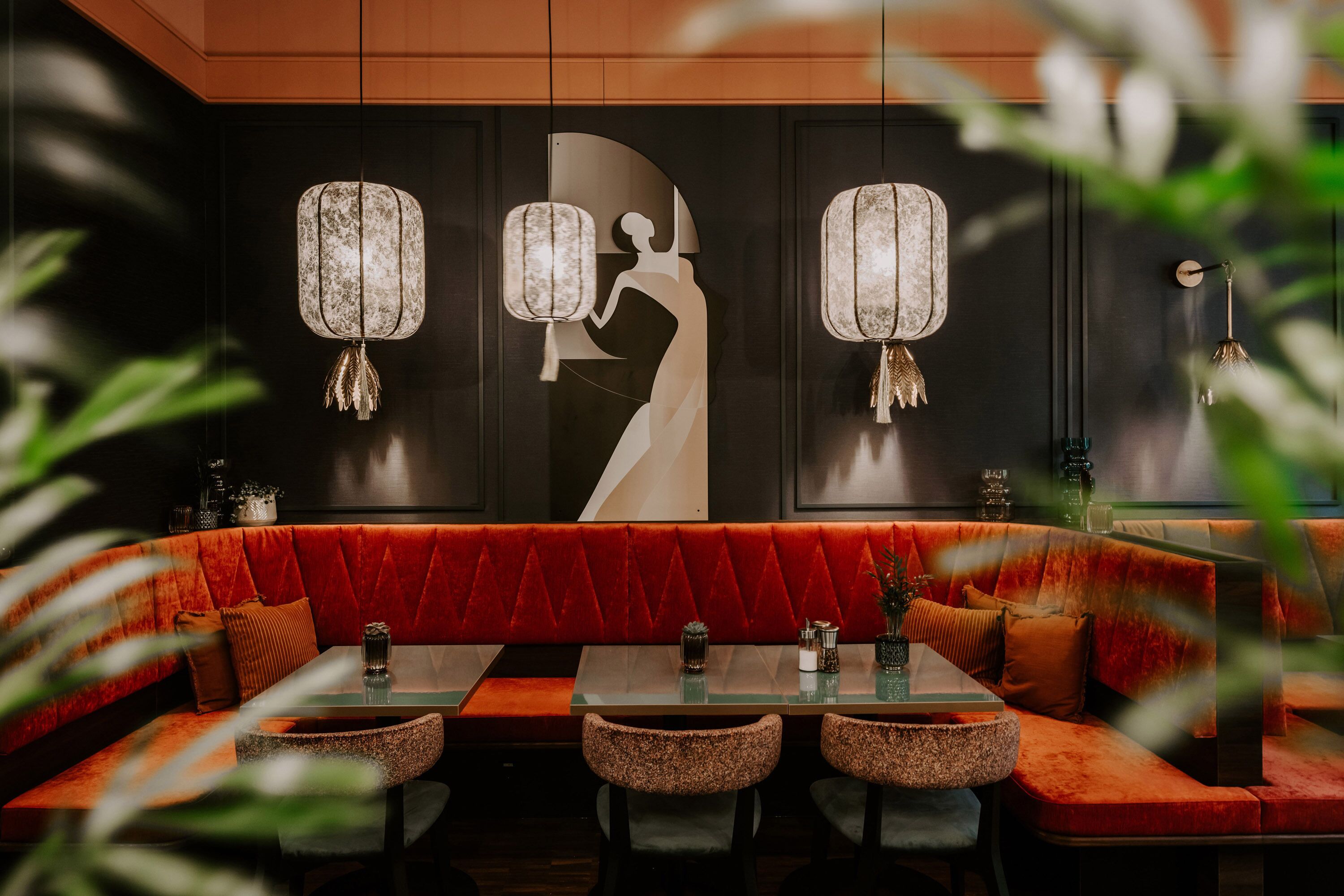A restaurant with a red upholstered booth seating area, a glass-top table, and hanging lantern-style lights.