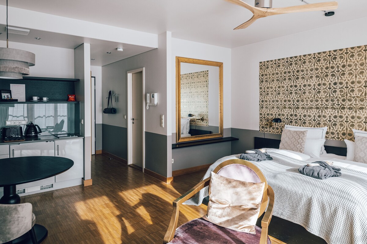A modern hotel room with a wooden floor, a dining area with a round table, a bed with a patterned headboard, and a large mirror.