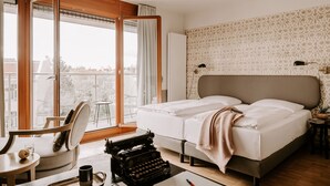 Hotellihuone, jossa on suuri sänky, kirjoituskoneella varustettu työpöytä, tuoli ja suuri ikkuna, josta avautuu näkymä kaupunkiin.