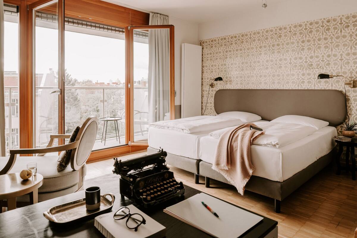 A hotel room with a large bed, a desk with a typewriter, a chair, and a view of the city through a large window.