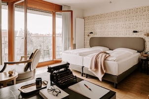 A hotel room with a large bed, a desk with a typewriter, a chair, and a view of the city through a large window.