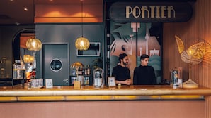 Baaritiskialue, jossa on kyltti, jossa lukee ”PORTIER”, ja kaksi henkilökunnan jäsentä. Siellä on riippuvalaisimia, tiski, jossa on erilaisia tuotteita, ja koristeellinen kasvi.
