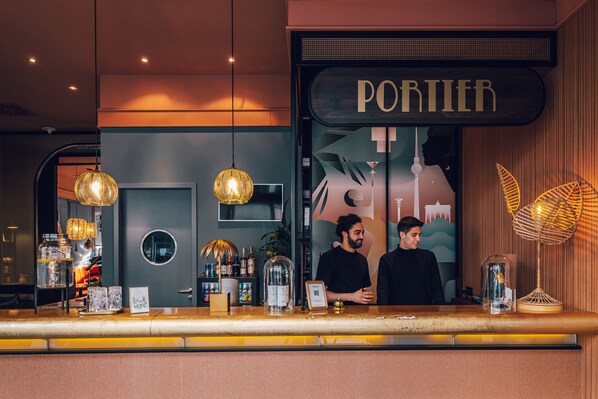 A bar area with a sign reading "PORTIER" and two staff members. There are hanging lamps, a counter with various items, and a decorative plant. - June Stay Berlin City West (Berlin)
