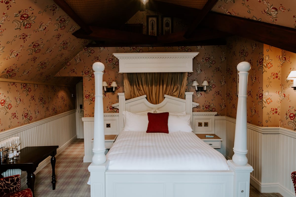 A four-poster bed with a red accent pillow, white headboard, and a floral wallpapered room.