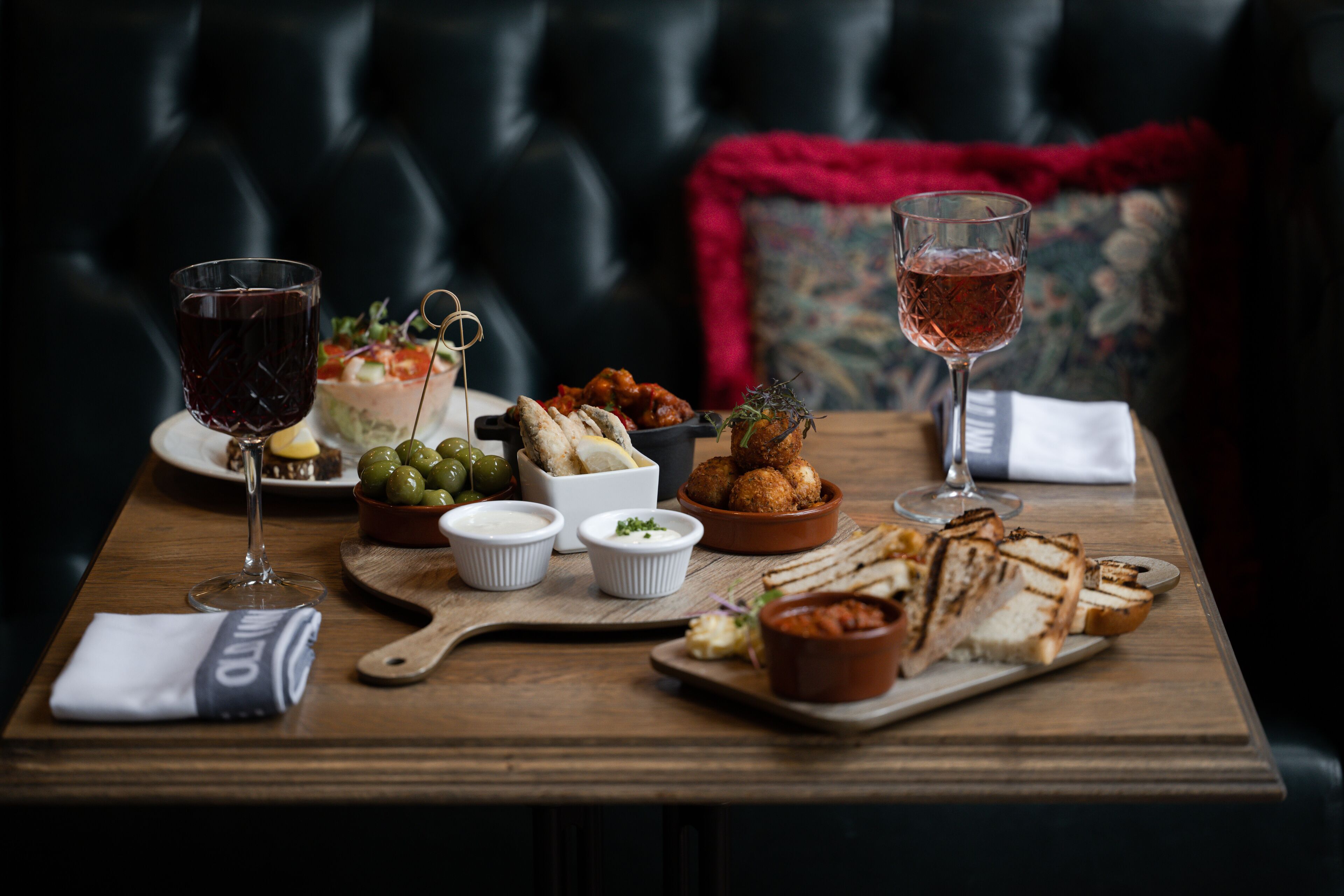 A dining table with various dishes and drinks, set against a dark upholstered sofa with a red throw blanket.