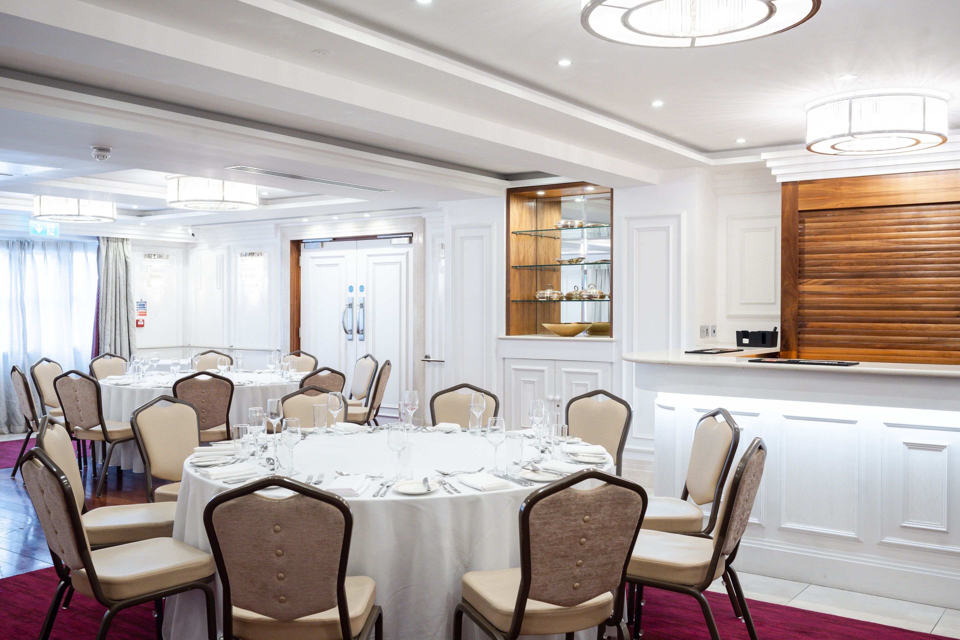 Uma sala de banquetes com mesas redondas preparadas para refeições, cadeiras e uma área de buffet com prateleiras de vidro.