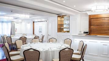 Uma sala de banquetes com mesas redondas preparadas para refeições, cadeiras e uma área de buffet com prateleiras de vidro.