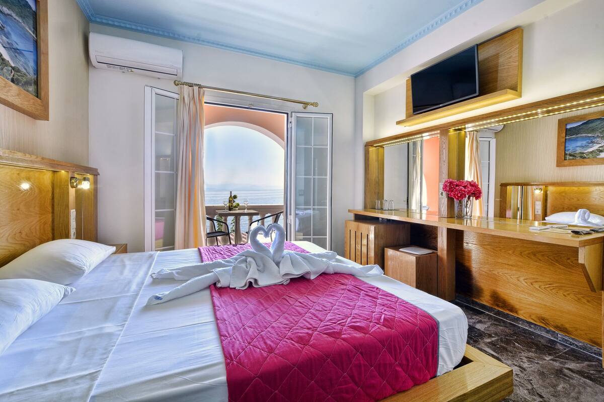 A hotel room with a bed, a desk, a TV, and a view of the sea.