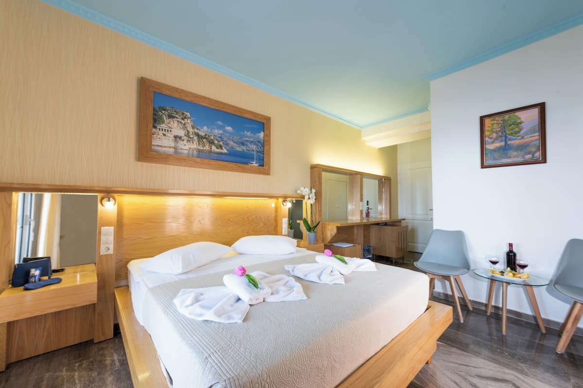 A hotel room with a bed, bedside tables, a desk, a chair, a small table with a bottle and glasses, and artwork on the walls.
