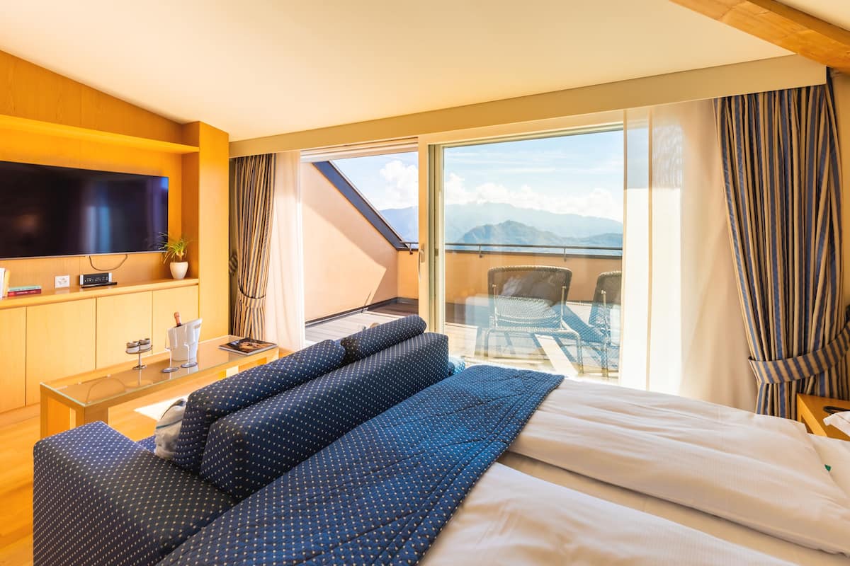 A hotel room with a bed, a sofa, a TV, a small table, and a balcony with a view of the mountains.