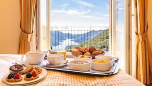 A breakfast tray with pastries, fruits, and beverages on a bed with a scenic view through a window.