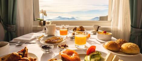 A breakfast table with various foods and drinks, a vase with flowers, and a view of mountains through a window.