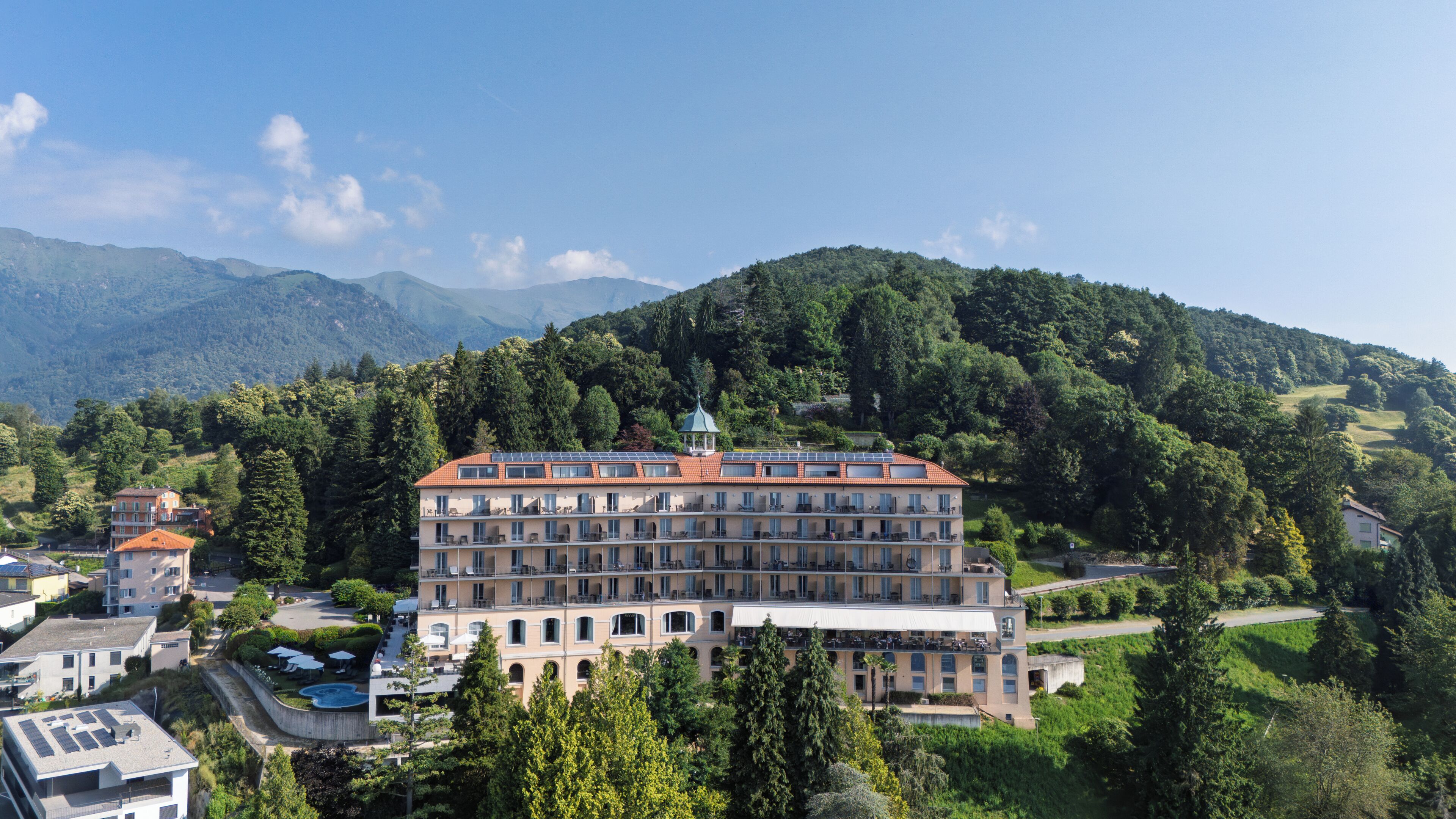 Ein mehrstöckiges Hotel mit rotem Ziegeldach, umgeben von üppiger Vegetation und Bergen im Hintergrund.