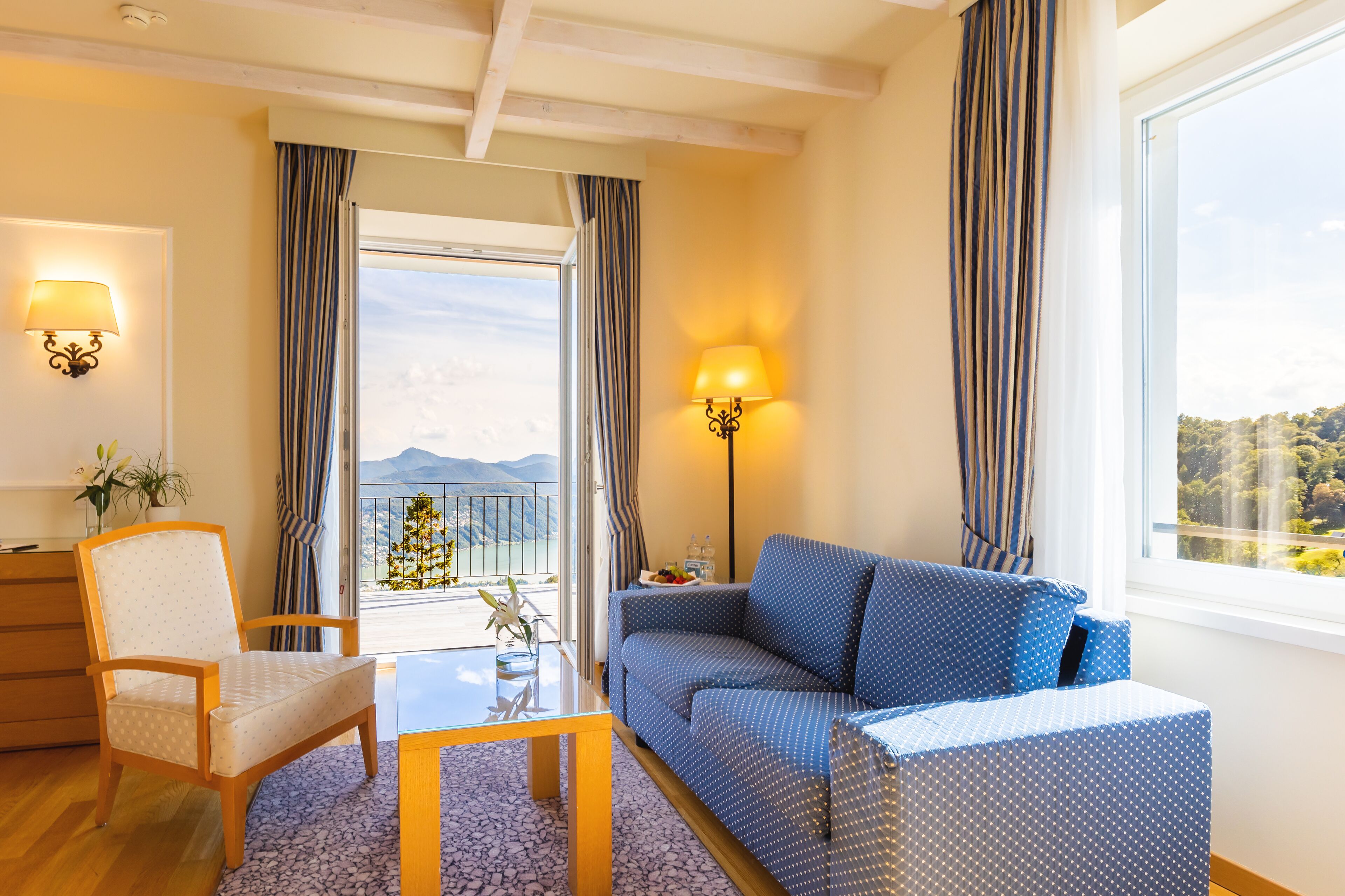 Habitación de hotel con sofá, sillón, mesa y balcón con cortinas.