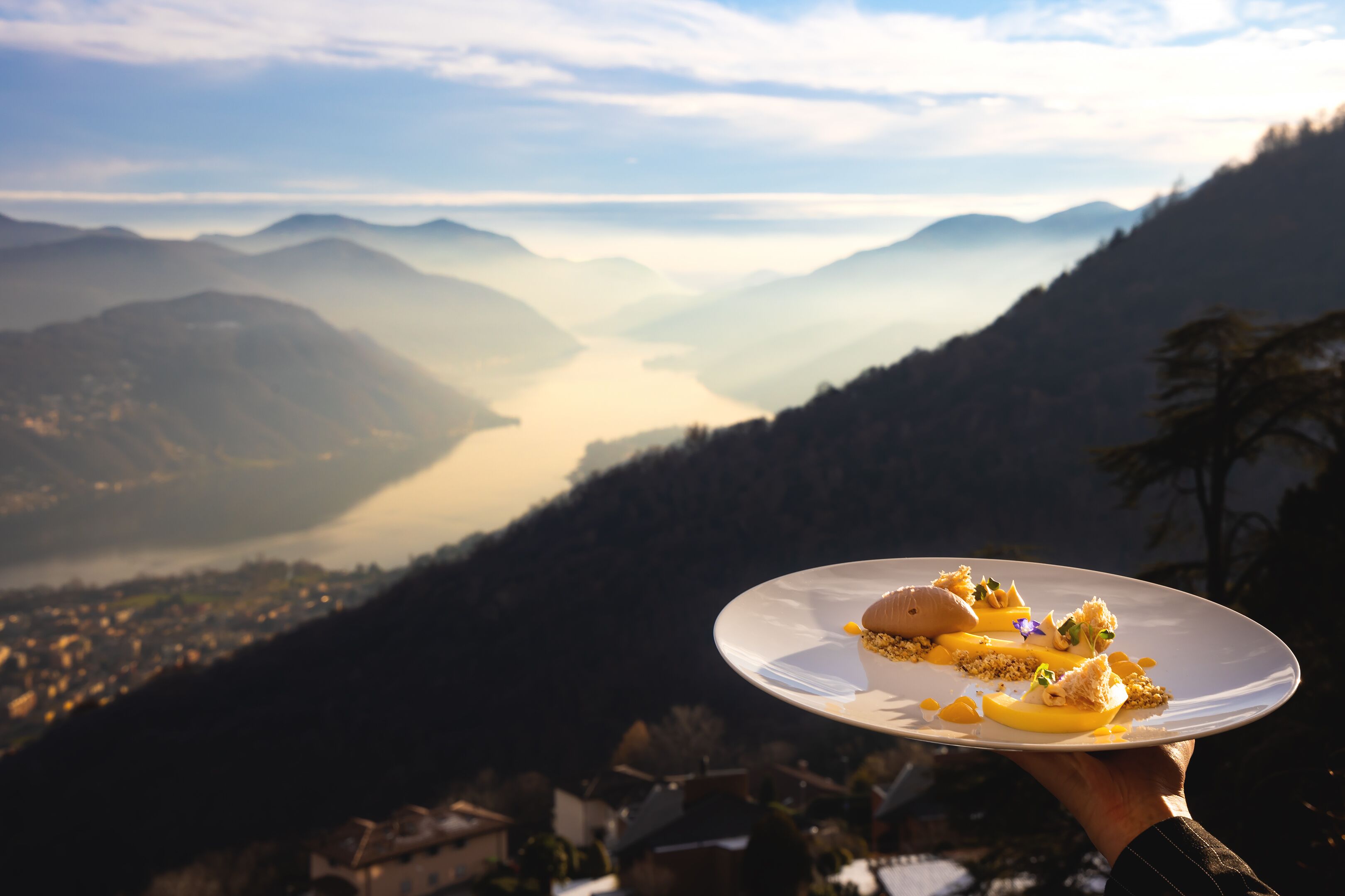 Una persona sosteniendo un plato de comida, con vistas a un paisaje montañoso que incluye un valle y un cuerpo de agua.