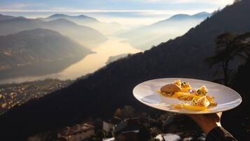 Una persona sosteniendo un plato de comida, con vistas a un paisaje montañoso que incluye un valle y un cuerpo de agua.