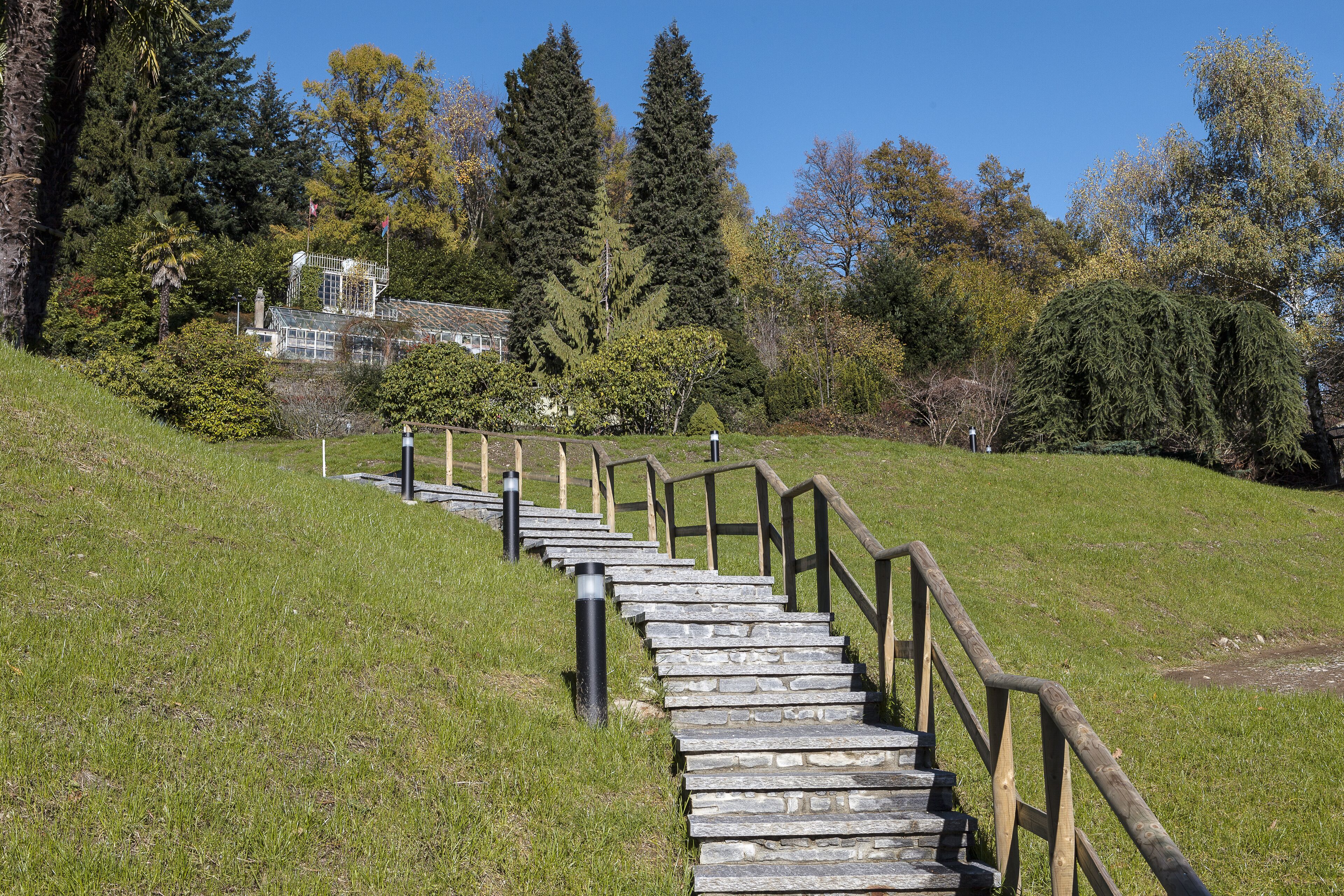 A stone staircase with wooden railings leading up a grassy slope, surrounded by trees and a building in the background.