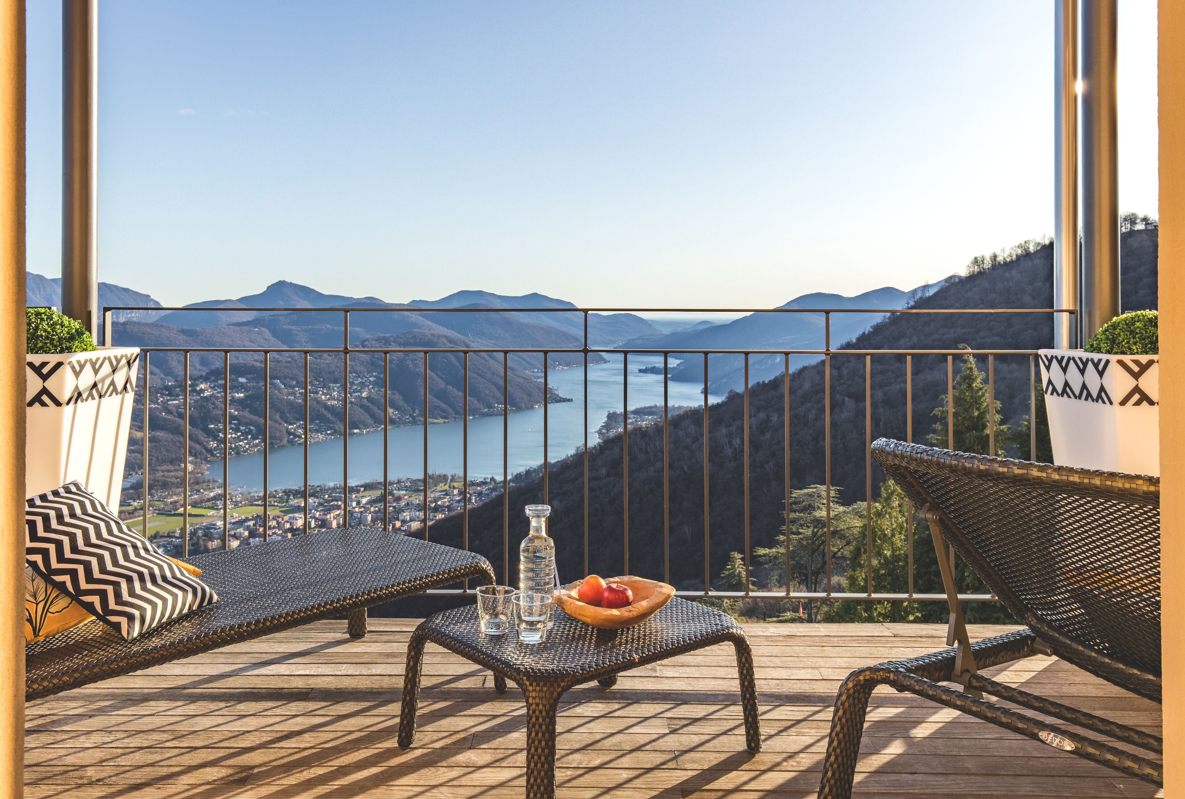 A balcony with wicker furniture, a table with a fruit bowl and glasses, and a scenic view of mountains and a lake.