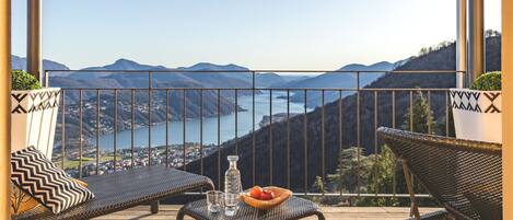Un balcón con mobiliario de mimbre, una mesa con frutero y vasos, y una vista panorámica de montañas y un lago.
