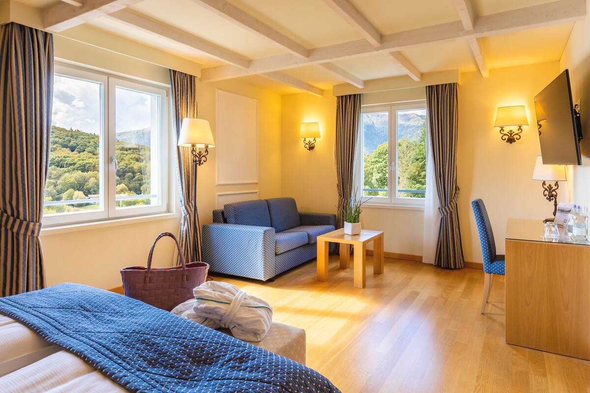 A hotel room with a bed, a sofa, a small table, and a TV. There are windows with curtains, and a view of greenery outside.