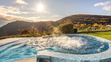 Una vasca idromassaggio con bordo piastrellato, circondata da un prato e con vista su un pendio boscoso.
