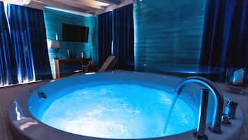 A large, round hot tub with a visible water flow, set in a room with a modern design, including a flat-screen TV and a seating area.