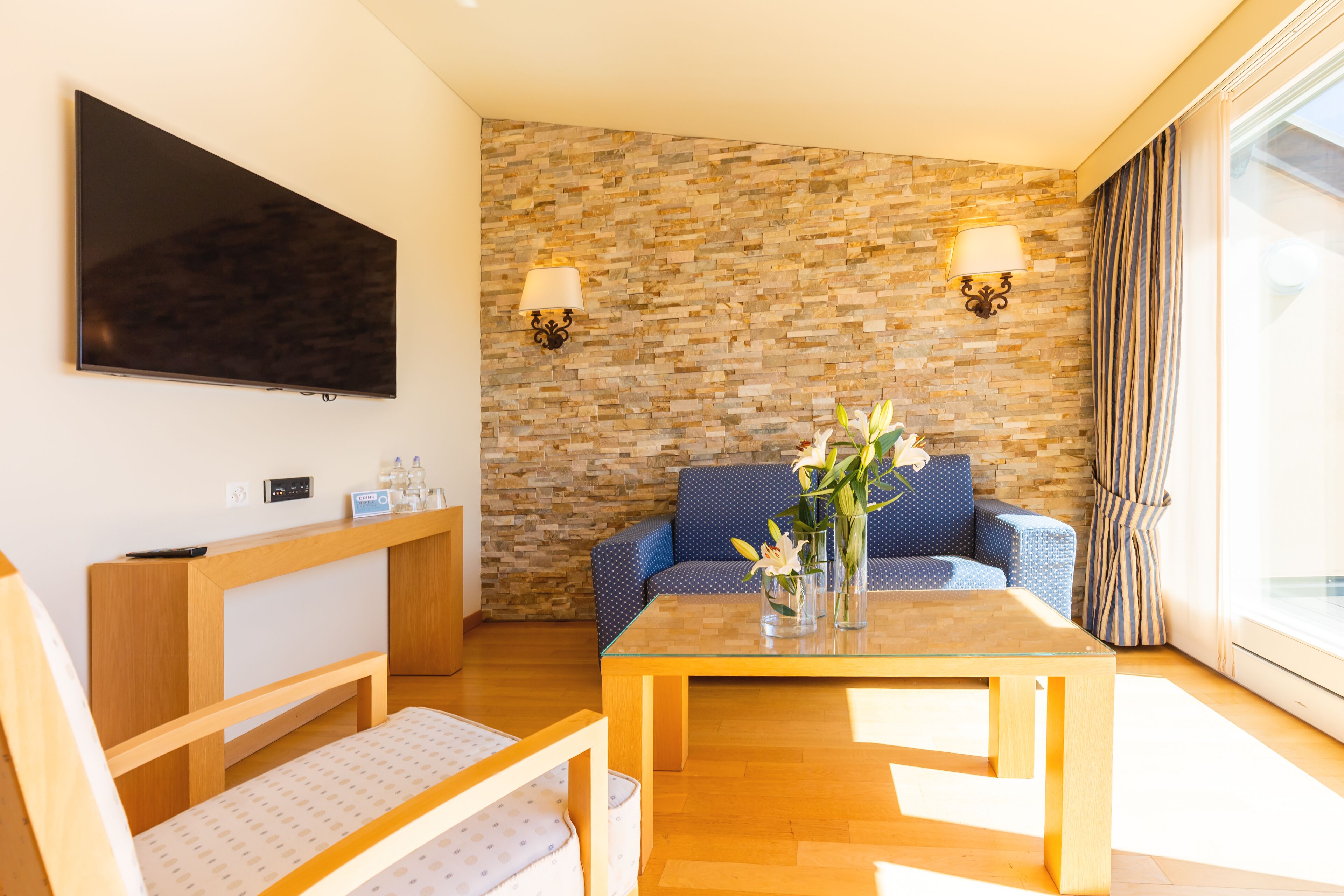 A modern hotel room with a stone wall, a flat-screen TV, a wooden desk, a blue sofa, and a glass coffee table.