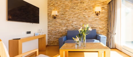 A modern hotel room with a stone wall, a flat-screen TV, a wooden desk, a blue sofa, and a glass coffee table.