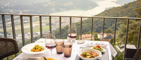 A dining table with plates of food, glasses of wine, and a scenic view of mountains and a lake.
