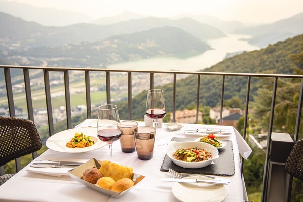 A dining table with plates of food, glasses of wine, and a scenic view of mountains and a lake. - Kurhaus Cademario Hotel & SPA (Cademario)