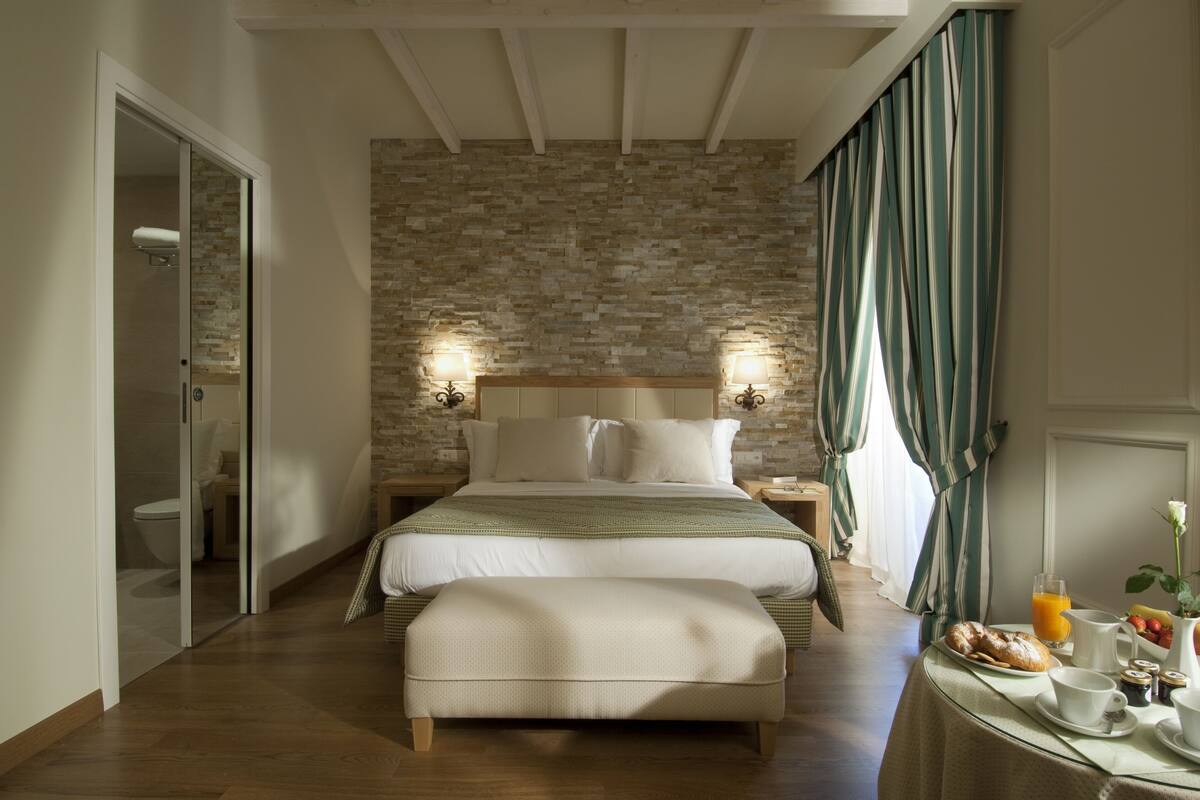 A bedroom with a stone wall, a bed, an ottoman, a nightstand with lamps, and a breakfast table with fruit and a vase.