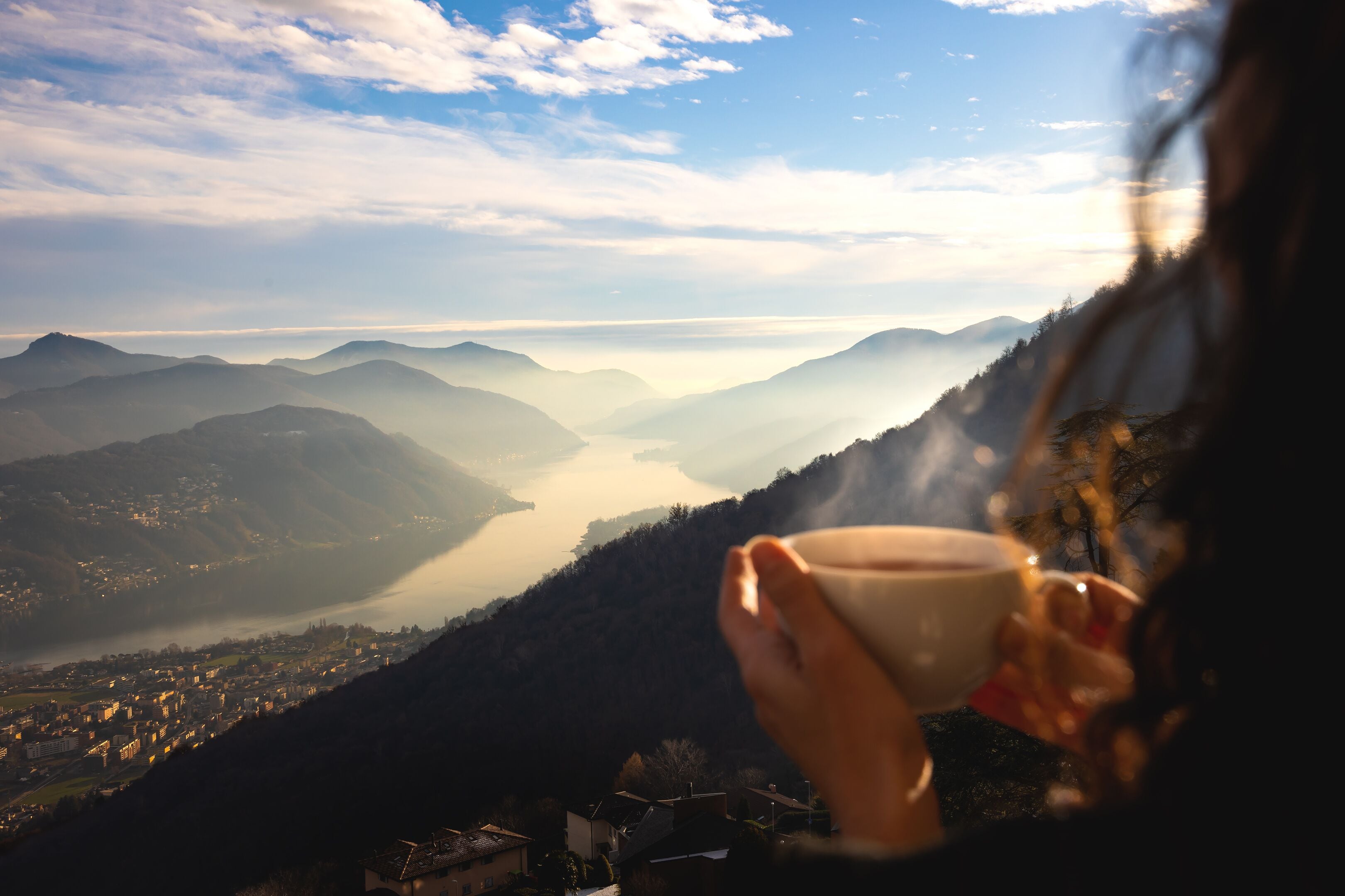 A person holding a cup, overlooking a valley with mountains and a body of water.
