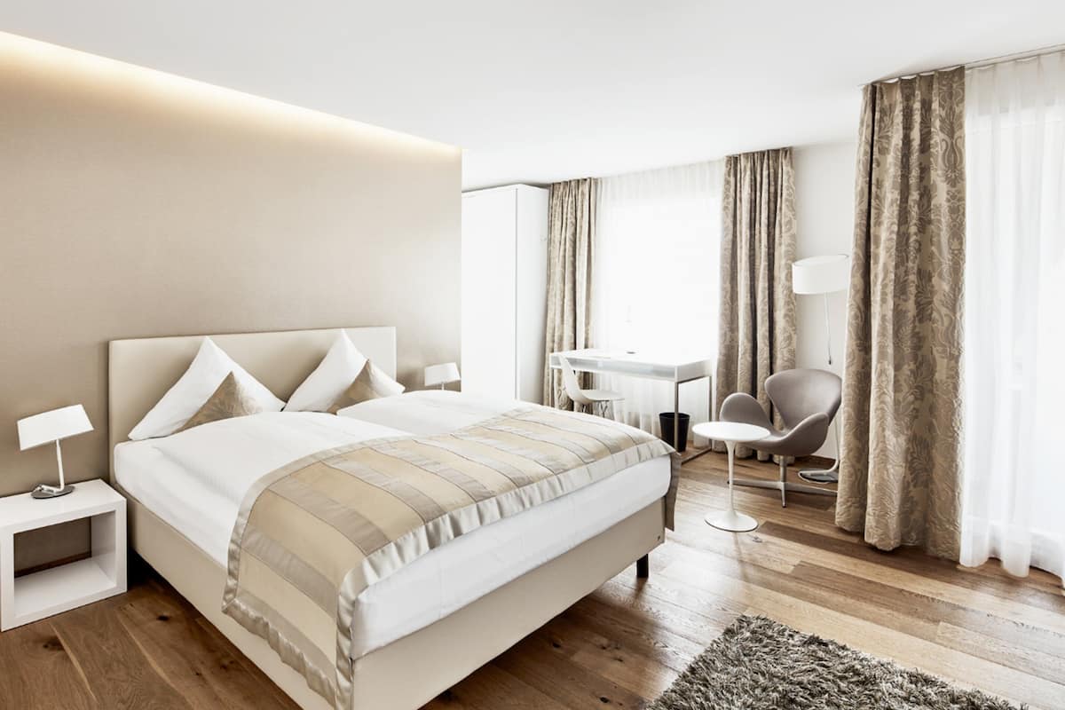 A modern hotel room with a large bed, bedside table, lamp, desk, chair, and curtains.