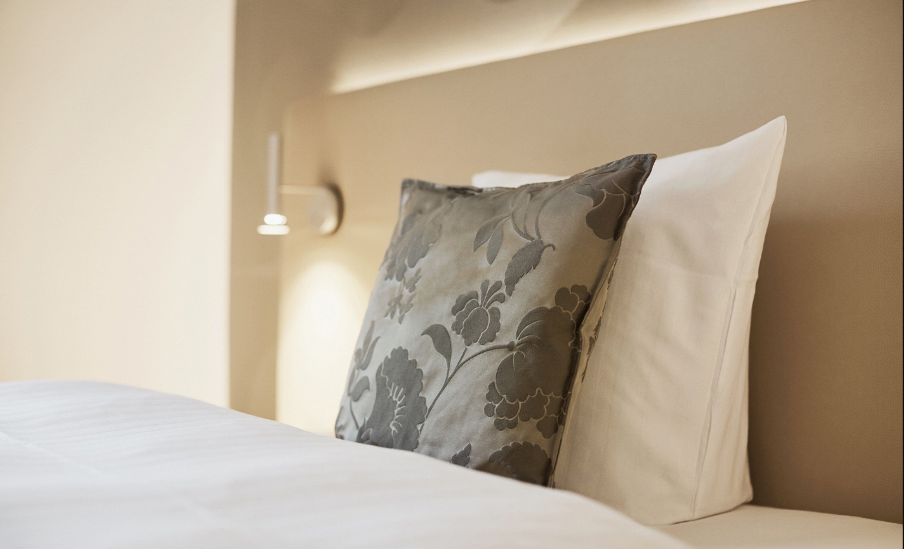 A bed with a floral pillow and a plain pillow, a wall-mounted light, and a plain wall.