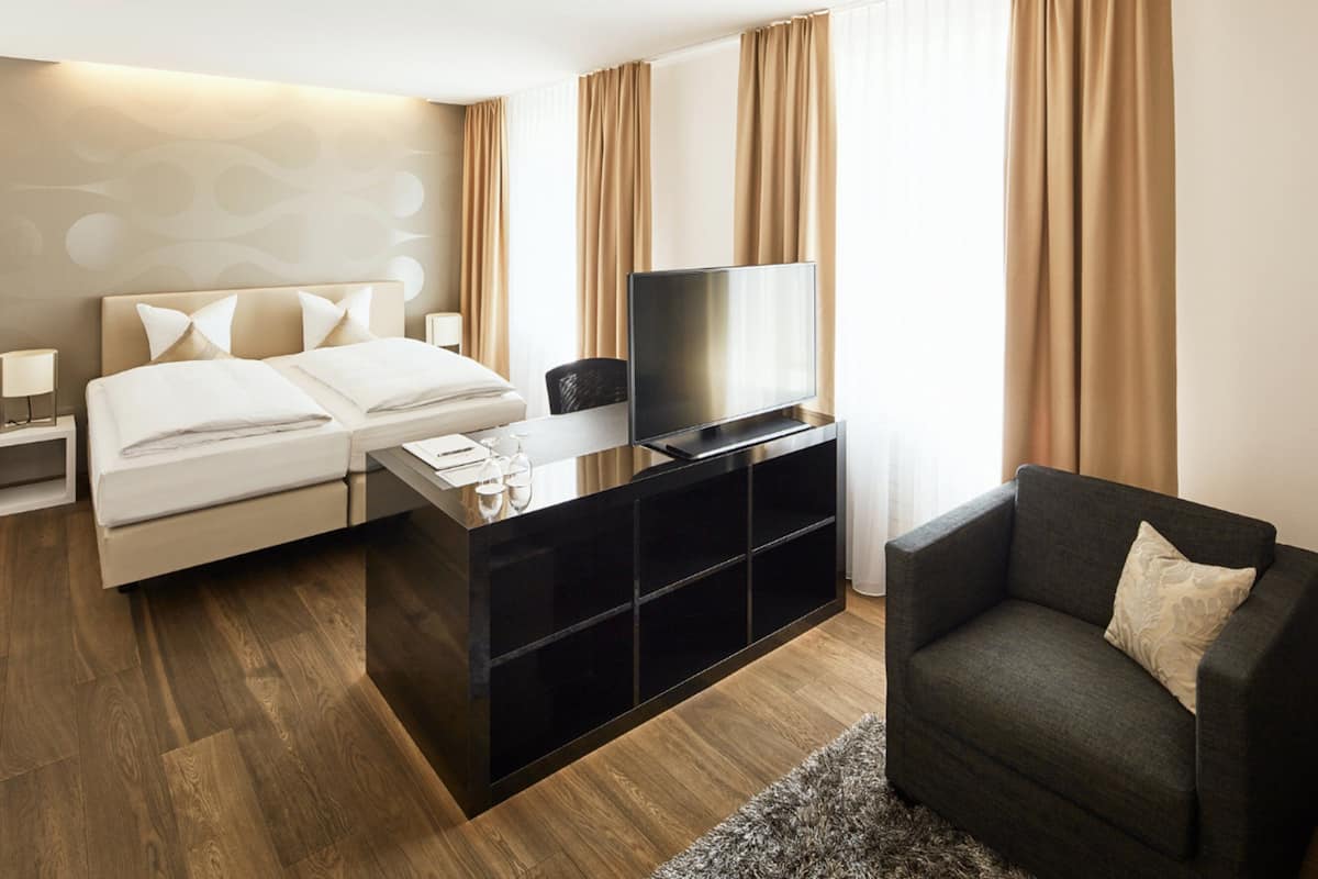 A modern hotel room with a bed, a TV on a black stand, a small table with a glass, and a grey armchair.