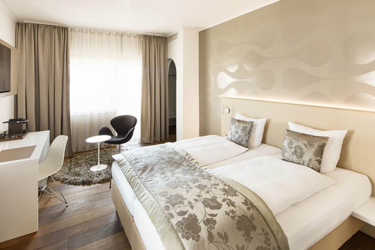 A hotel room with a bed, a desk, a chair, and a view of the outdoors through large windows.