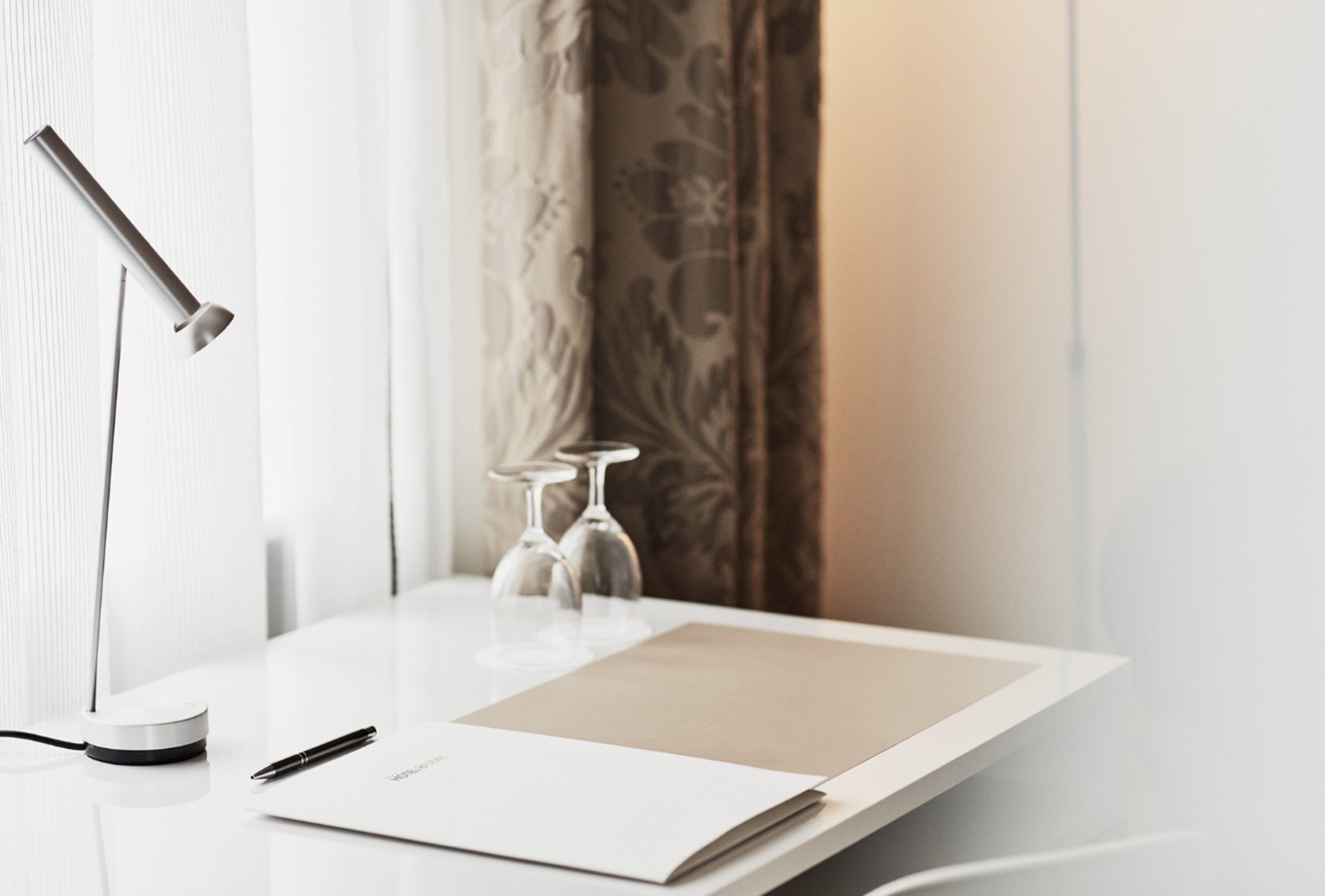 A modern desk with a white surface, a pen, a notebook, a lamp, and two wine glasses.